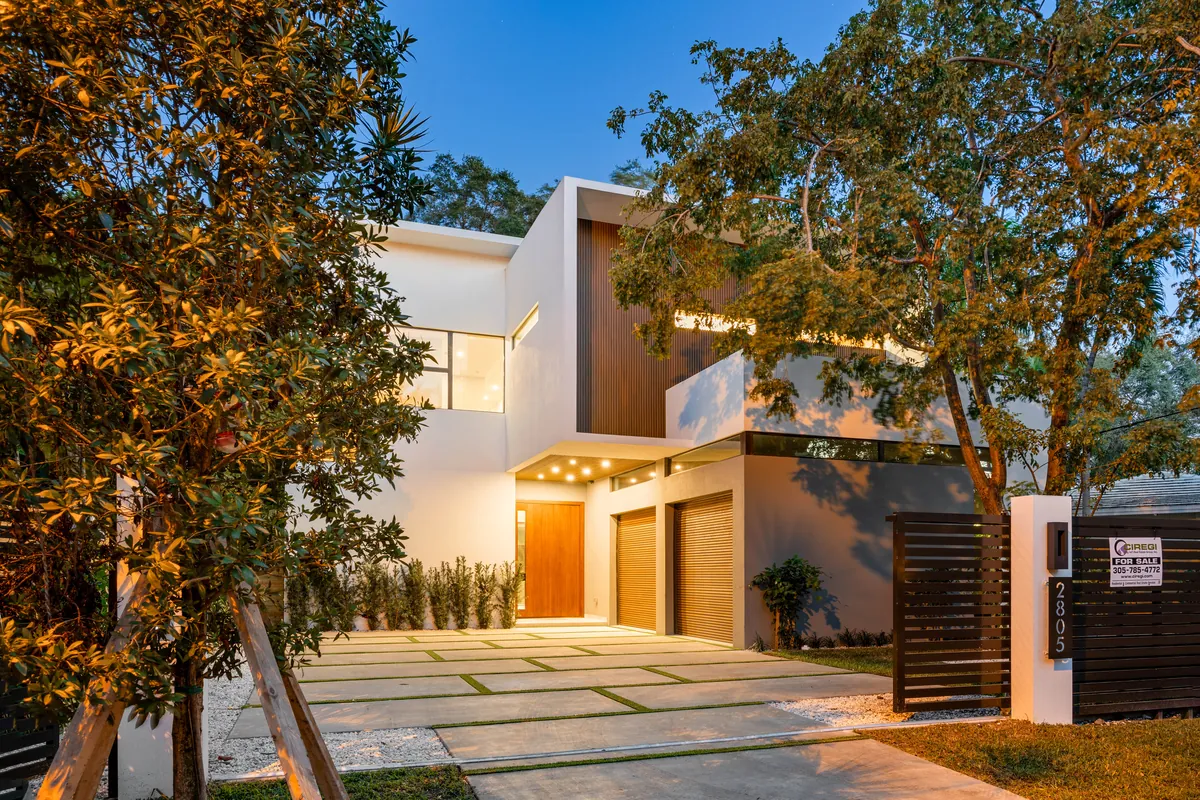 Modern white home with wood accents, an illuminated entrance and trees at dusk