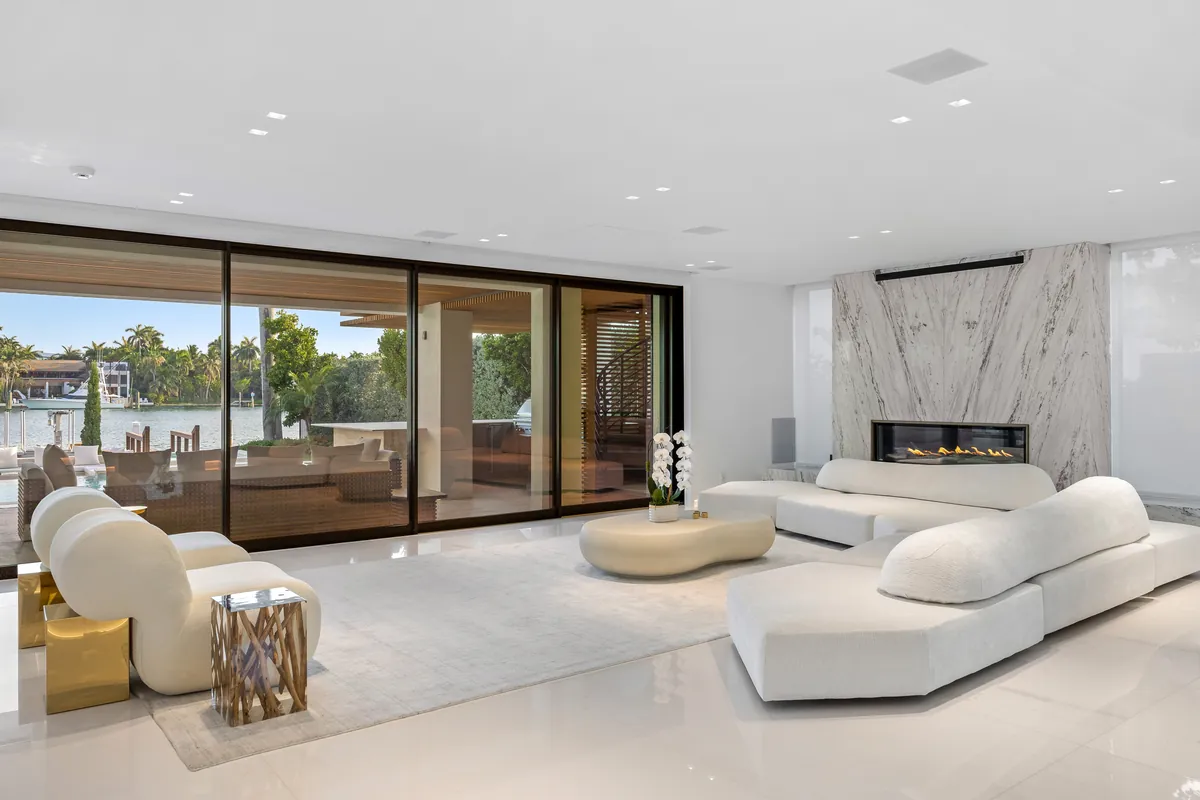 Pale curved sofas beside a stone fireplace and open glass doors to a waterfront terrace