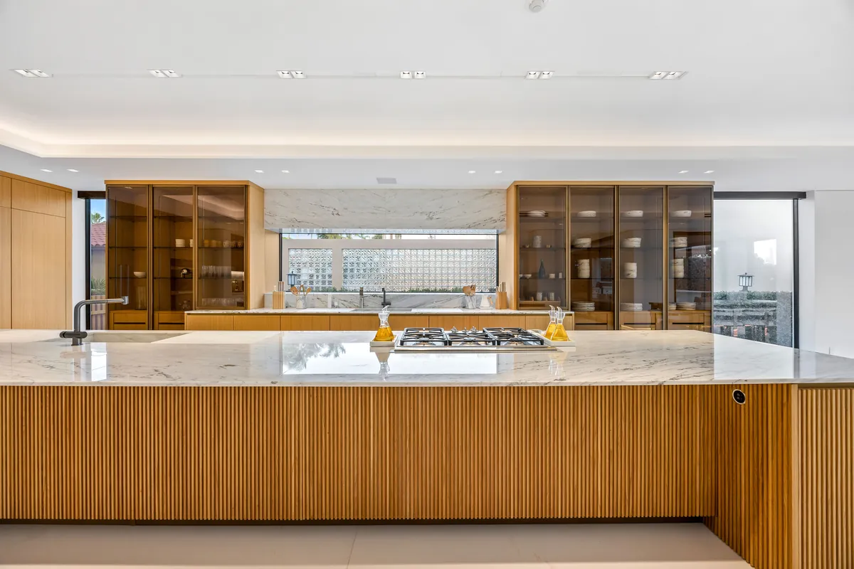Long kitchen island with wood cabinetry, pale stone and glass-fronted cupboards