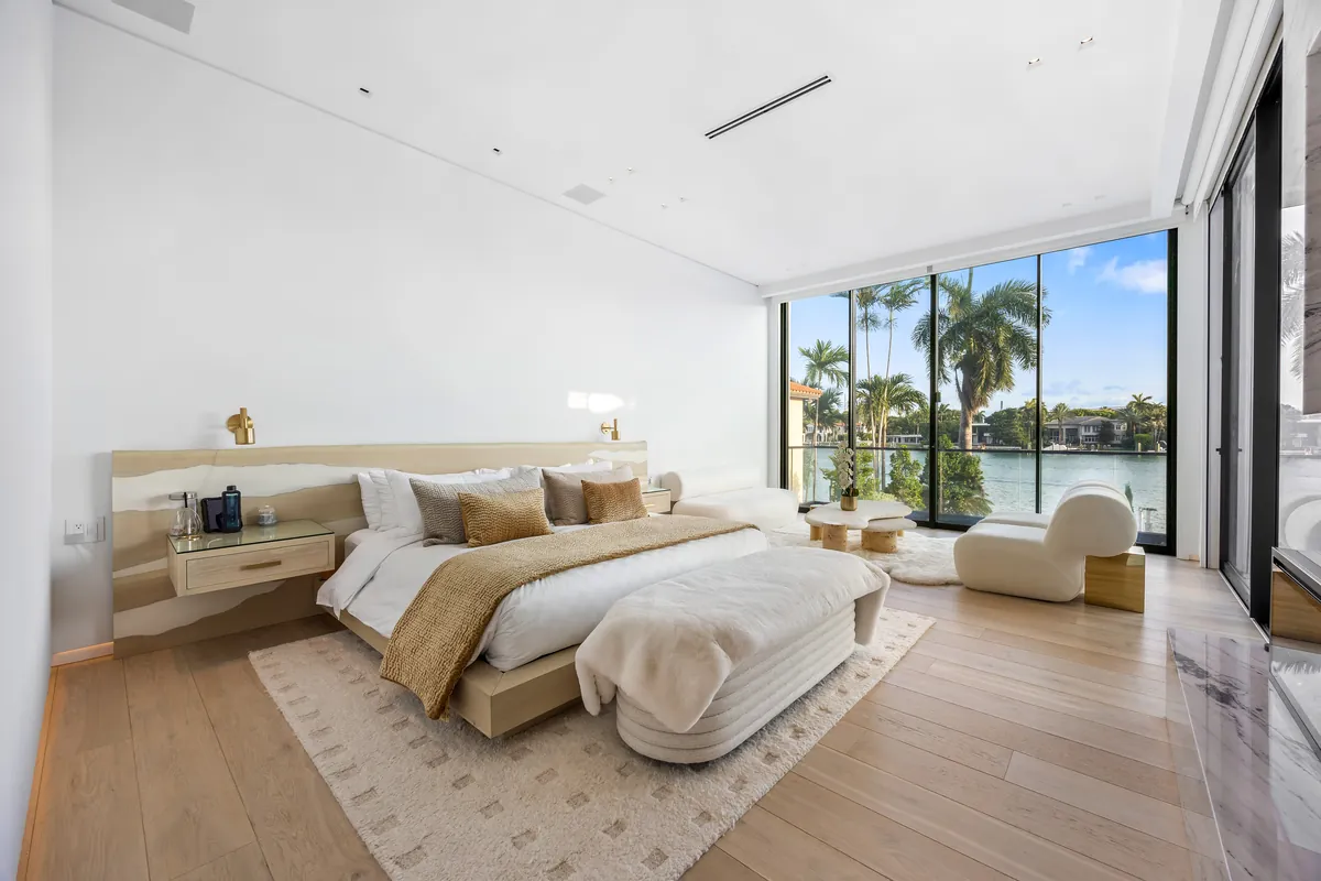 Bedroom with a low upholstered bed, pale seating and tall windows facing the water