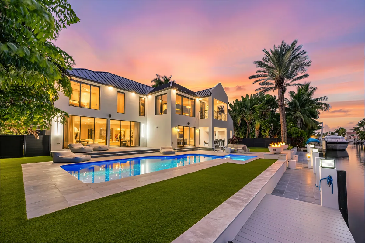 Illuminated waterfront home with a long pool, palms and a pink evening sky