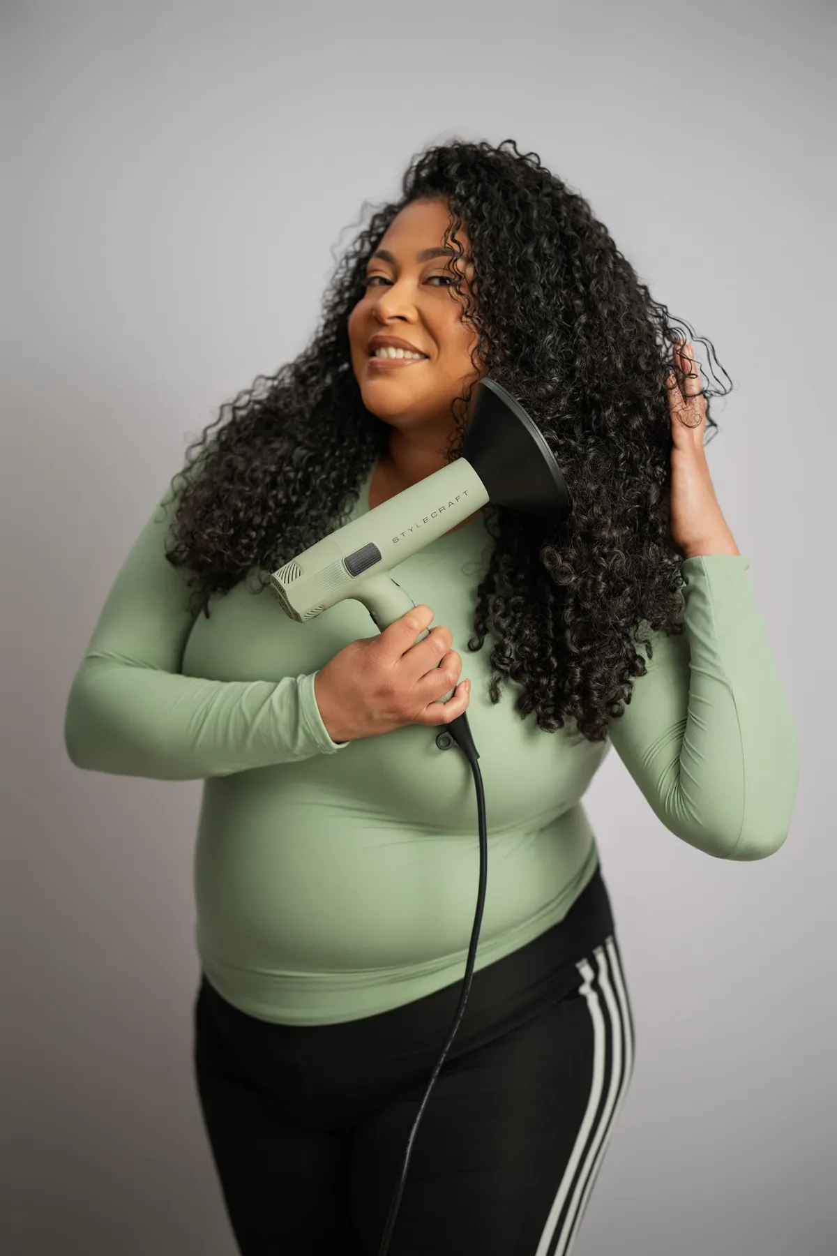 Model with curly hair holding a green Stylecraft hair dryer in a studio portrait