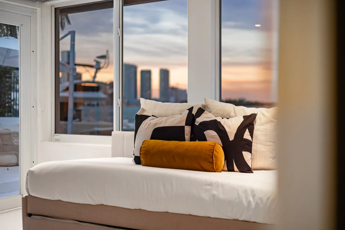 Pale daybed with patterned cushions beside waterfront windows at sunset